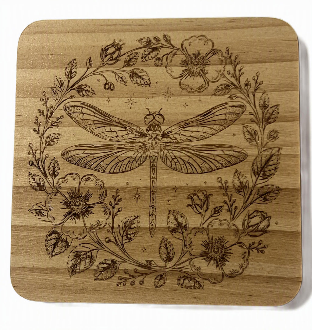 A square wooden coaster with rounded corners, featuring a laser-engraved design of a dragonfly surrounded by a floral wreath.