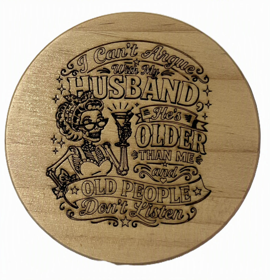 A round wooden coaster with a laser-engraved design featuring a skeleton woman holding a martini glass and humorous text about husbands and old people.