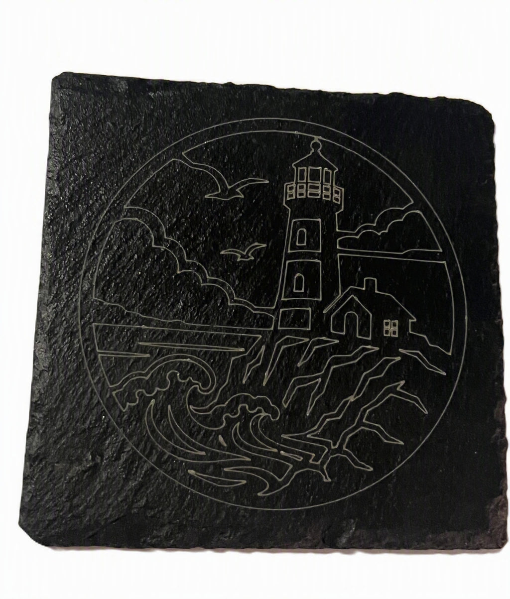 A square black slate coast sign featuring a white line art design of a lighthouse, waves, birds, and a small house.
