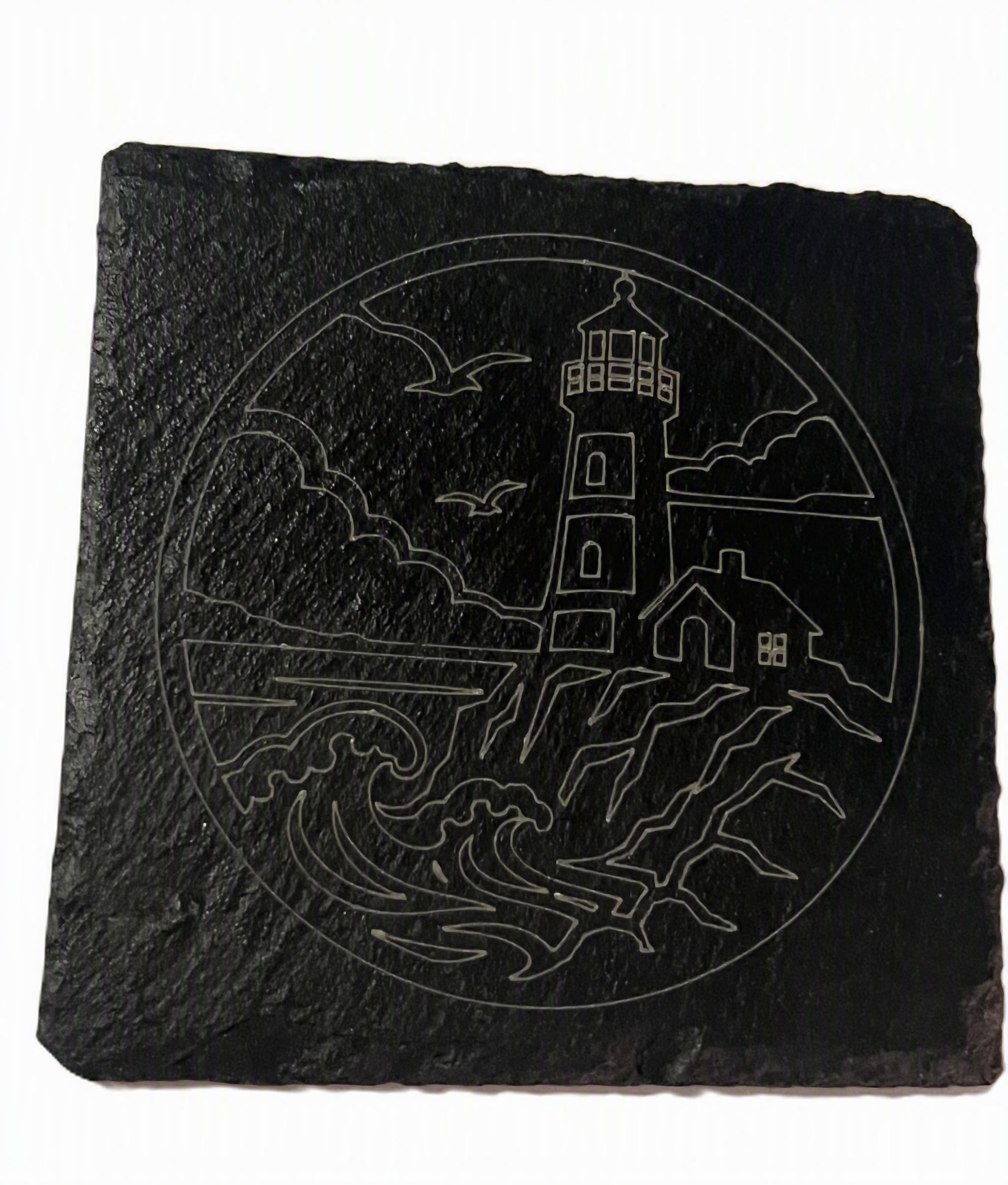 A square black slate coast sign featuring a white line art design of a lighthouse, waves, birds, and a small house.