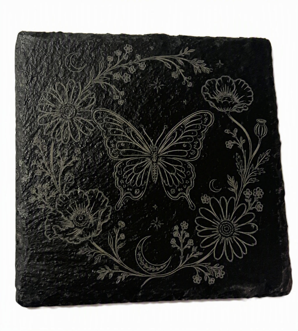 A square black slate coaster featuring a white engraved design of a butterfly, flowers, and a crescent moon, shown on a plain white background.