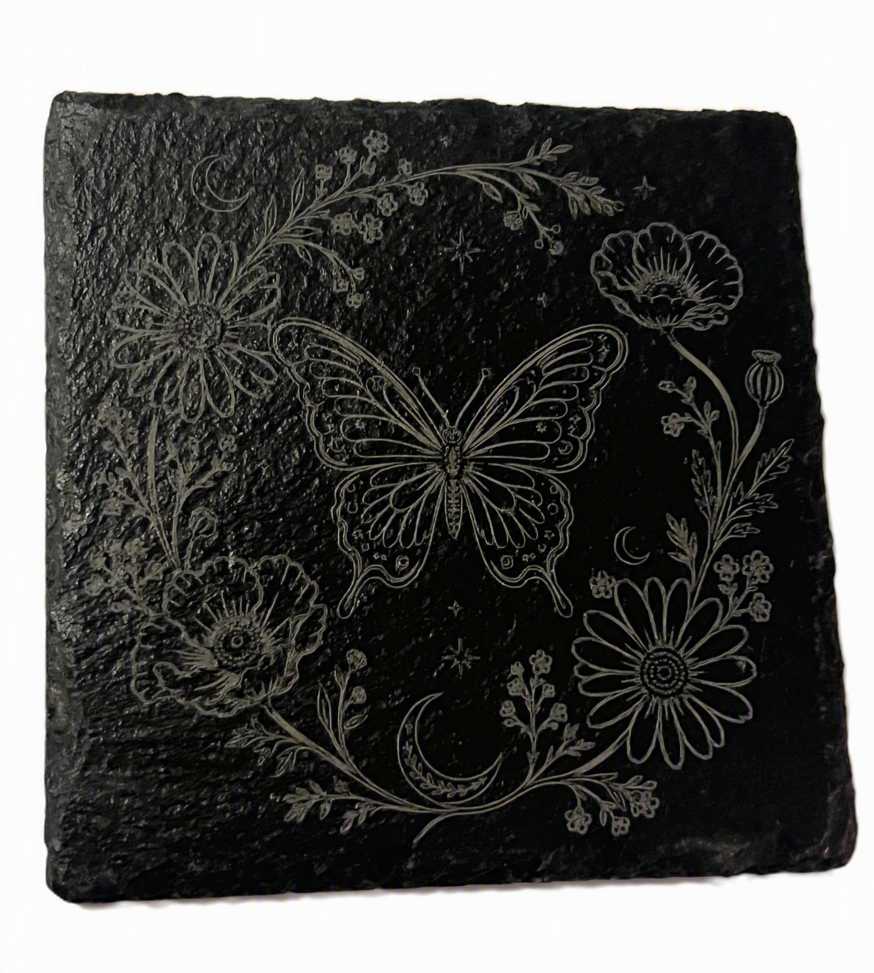 A square black slate coaster featuring a white engraved design of a butterfly, flowers, and a crescent moon, shown on a plain white background.