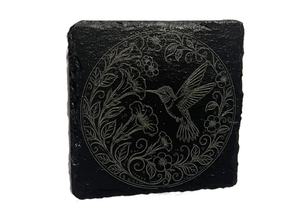 A square black slate coaster featuring an engraved circular design of a hummingbird and floral elements.