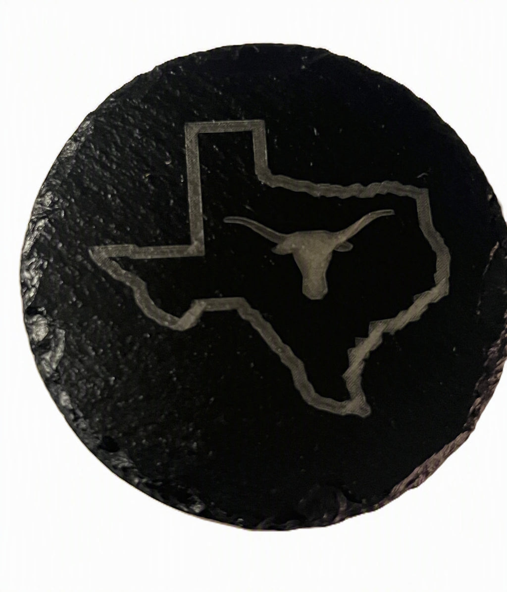 A round black slate coaster featuring an engraved outline of the state of Texas with a longhorn bull silhouette in the center.