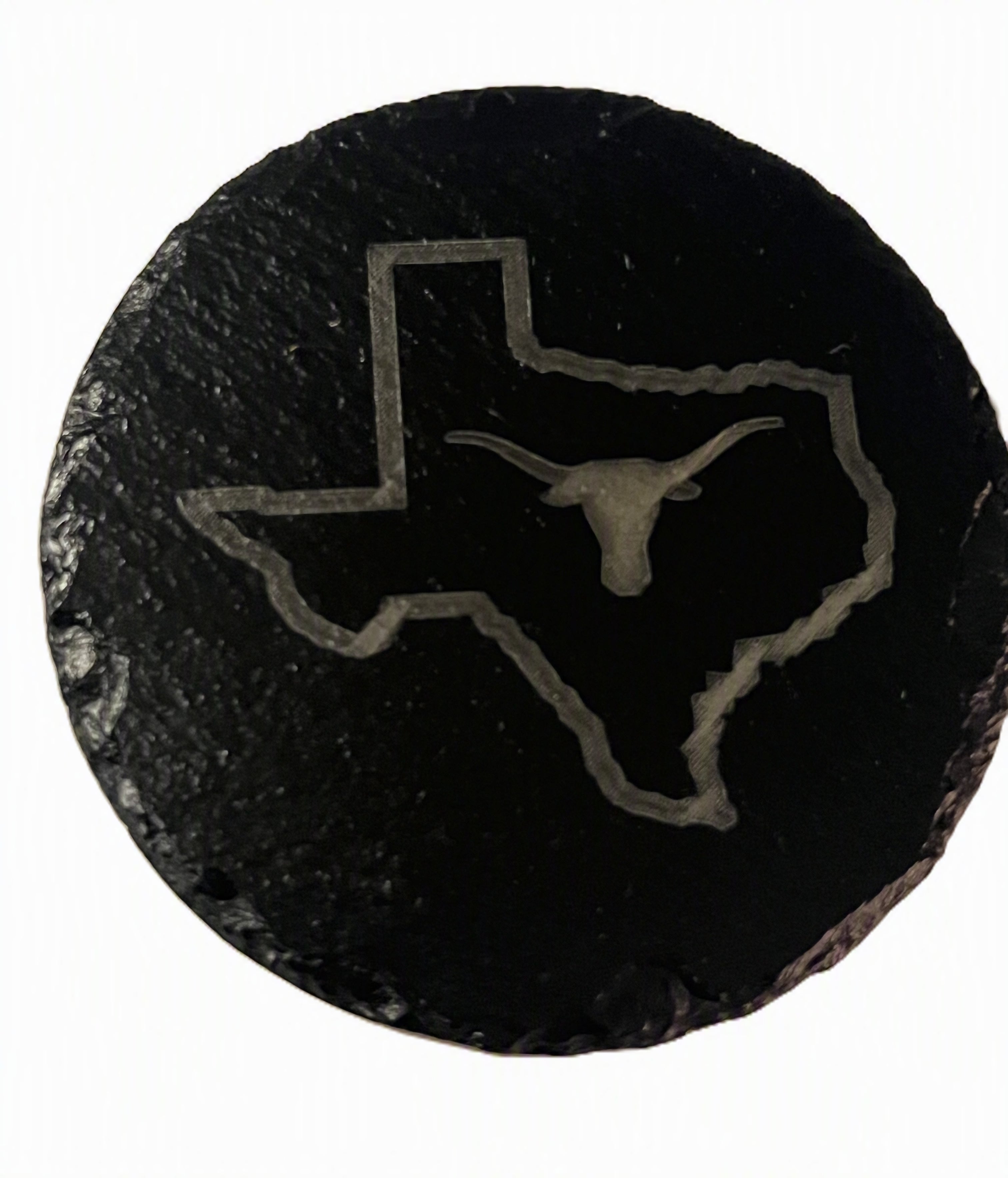 A round black slate coaster featuring an engraved outline of the state of Texas with a longhorn bull silhouette in the center.
